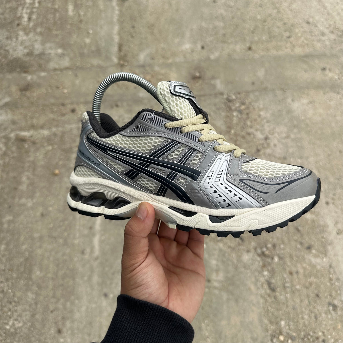 A silver and gray Asics running shoe held up by a hand against a concrete wall background.