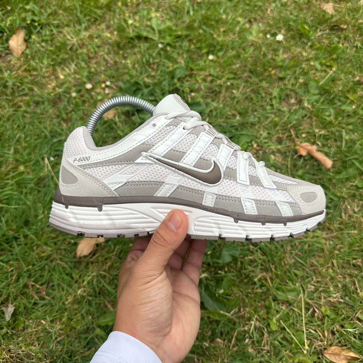 A Nike P-6000 sneaker in light gray and white, held by a hand against a background of green grass.