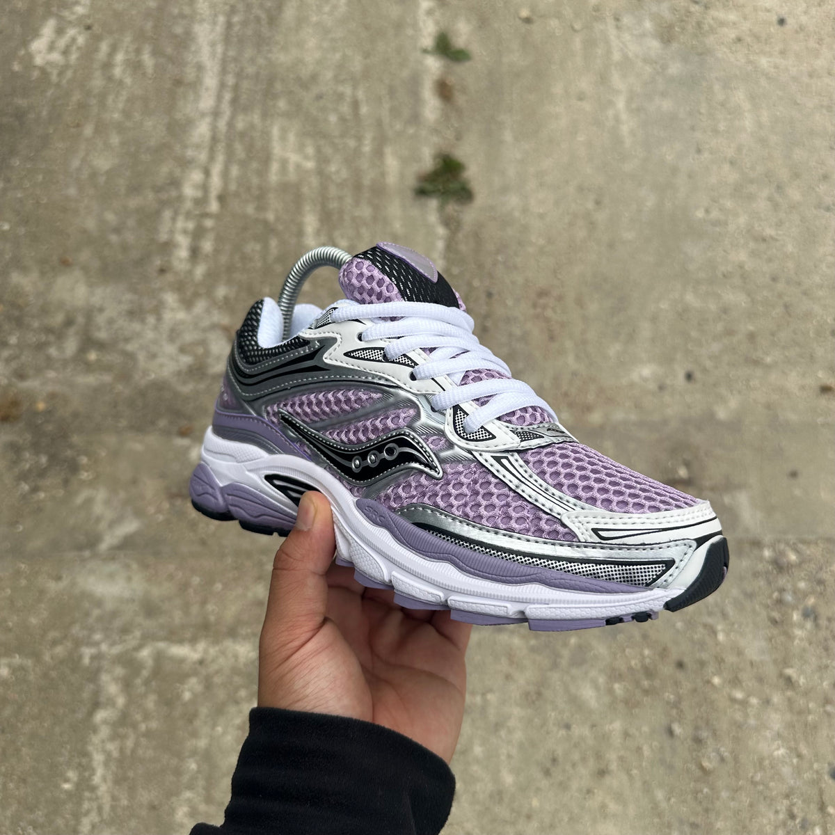 A hand holding a Saucony running shoe in purple, white, and black, photographed against a concrete ground background.