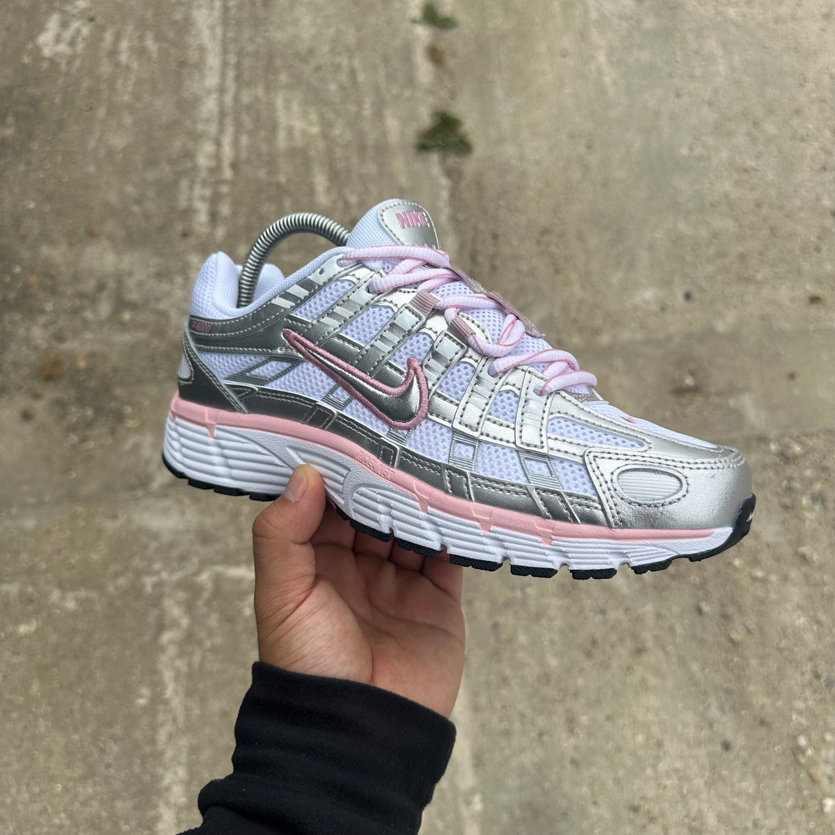 A Nike running shoe in metallic silver, white, and pink, held up by a hand against a concrete wall background.