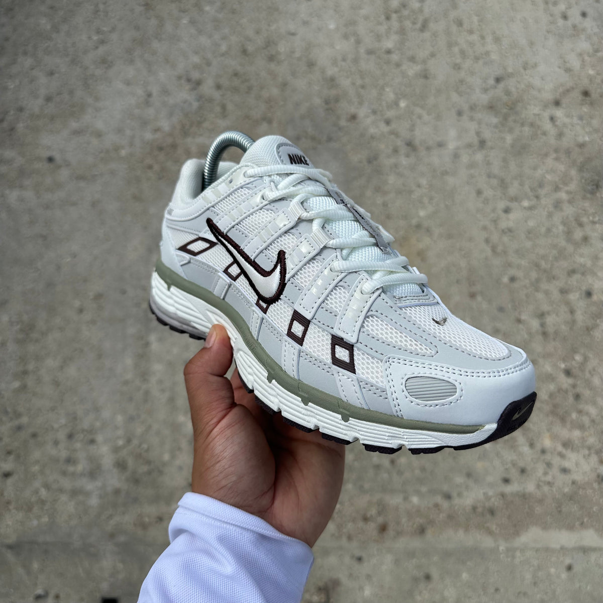 A white Nike sneaker with black and olive green accents, held up by a hand against a concrete wall background.